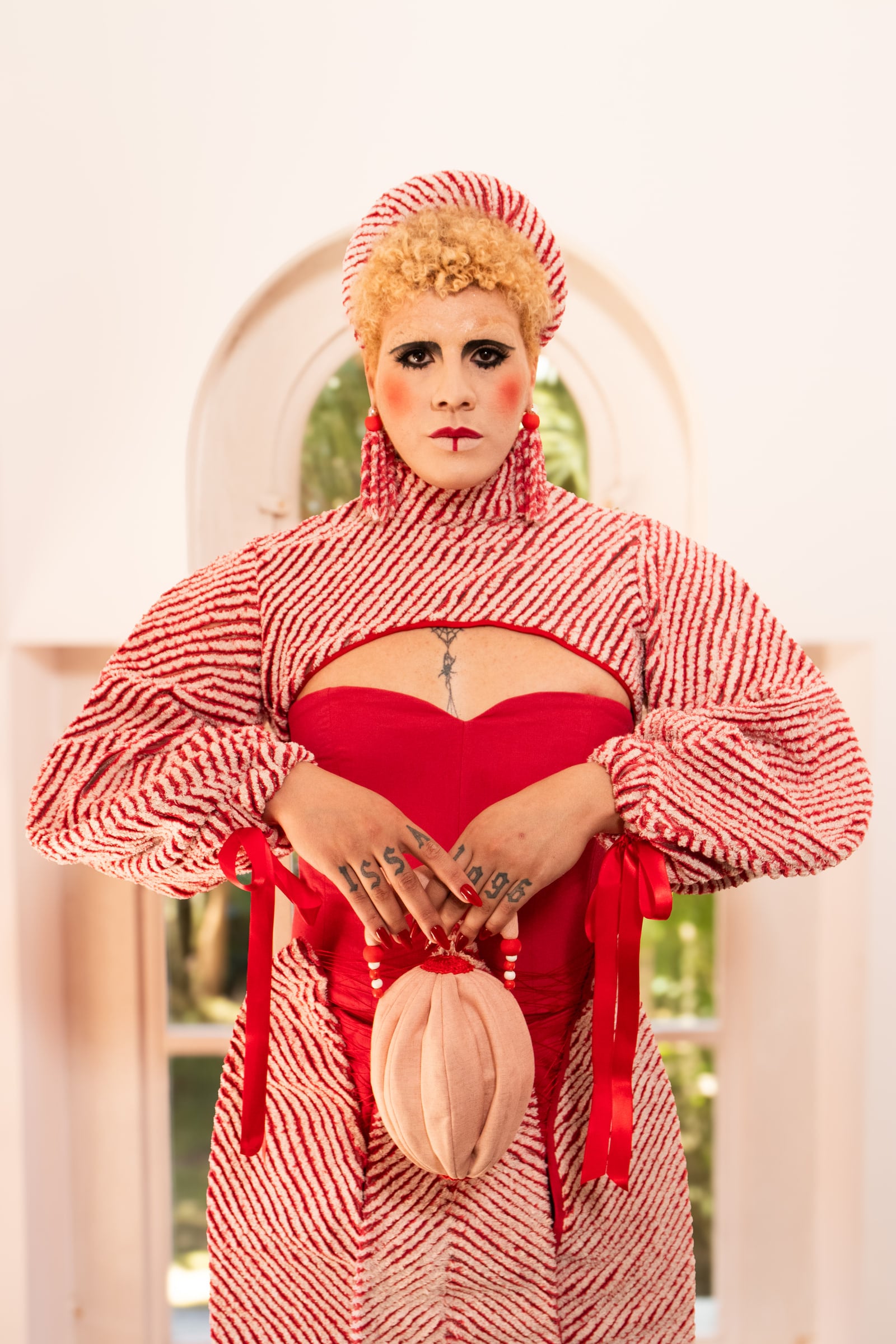 Model facing the camera before an arched window, hands resting on a pale oval accessory, the red dress beneath the striped layer.